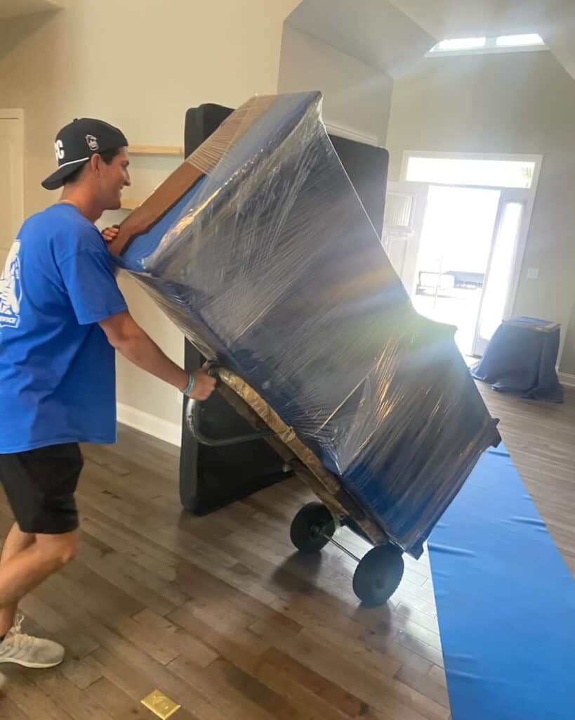 Professional mover transporting a fully wrapped piano using a dolly, showcasing careful handling and expert moving services for heavy and delicate items